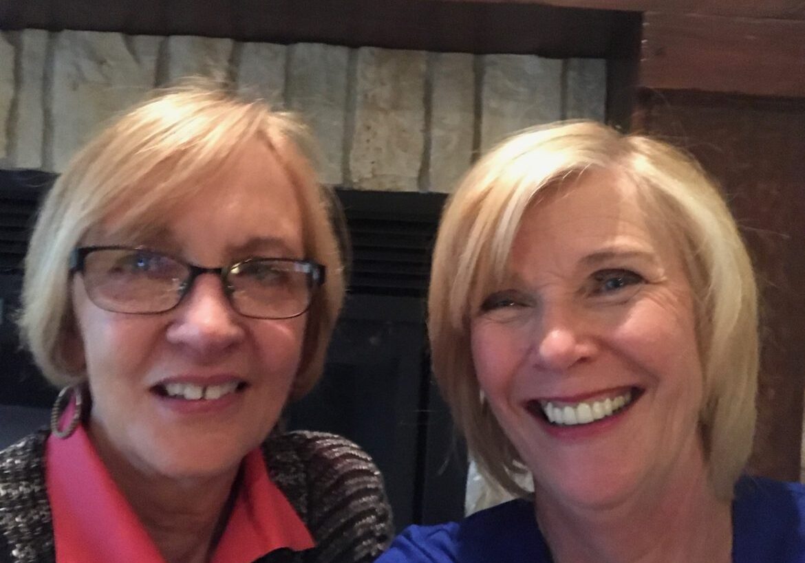 Two women smiling together indoors, one wearing glasses.